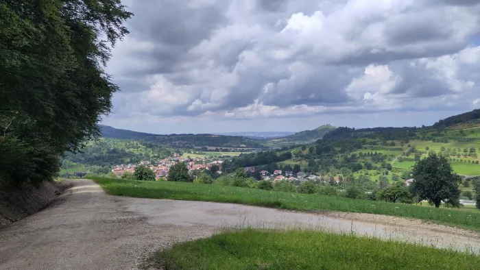 Ein Schotterweg führt links im Bild einen starken Abhang hinunter. Im Hintergrund offenbart sich ein Blick auf eine Stadt und ein paar Berge.