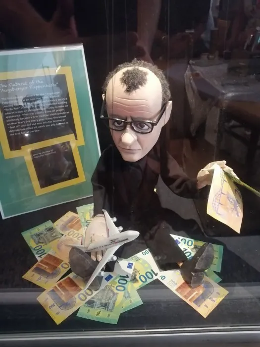 Eine Marionette von Friedrich Merz in der Augsburger Puppenkiste. Er ist umgeben von Geld und hat ein Spielzeugflugzeug an der Hand.