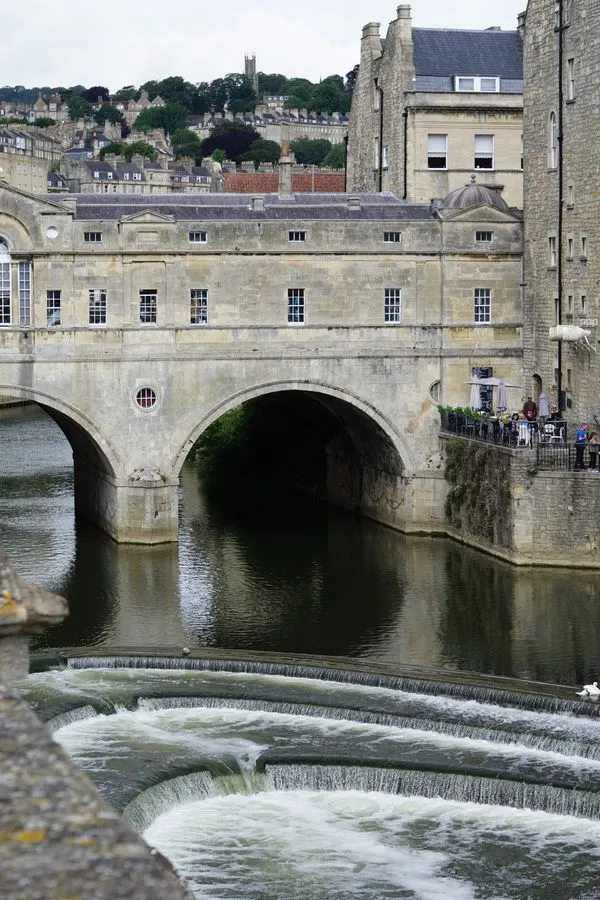 Der Avon Fluss fließt unter einer bebauten Brücke in der Innenstadt von Bath. Die Gebäude bestehen aus hellgrauen Stein. Im Hintergrund erstreckt sich die Stadt auf weiteren Hügeln.