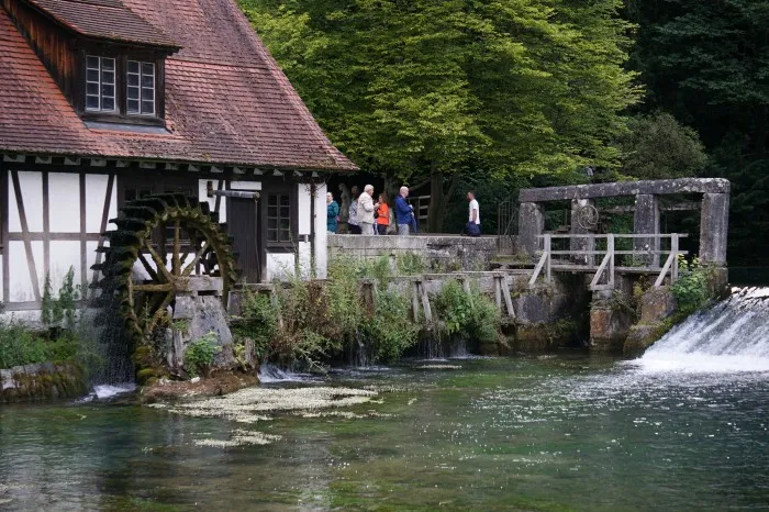 Der Blautopf. Ein kleine See, an dem der Fluss mit blauem Wasser entspring. Ein altes Fachwerkhaus mit Mühlrad ist ebenfalls zu sehen.