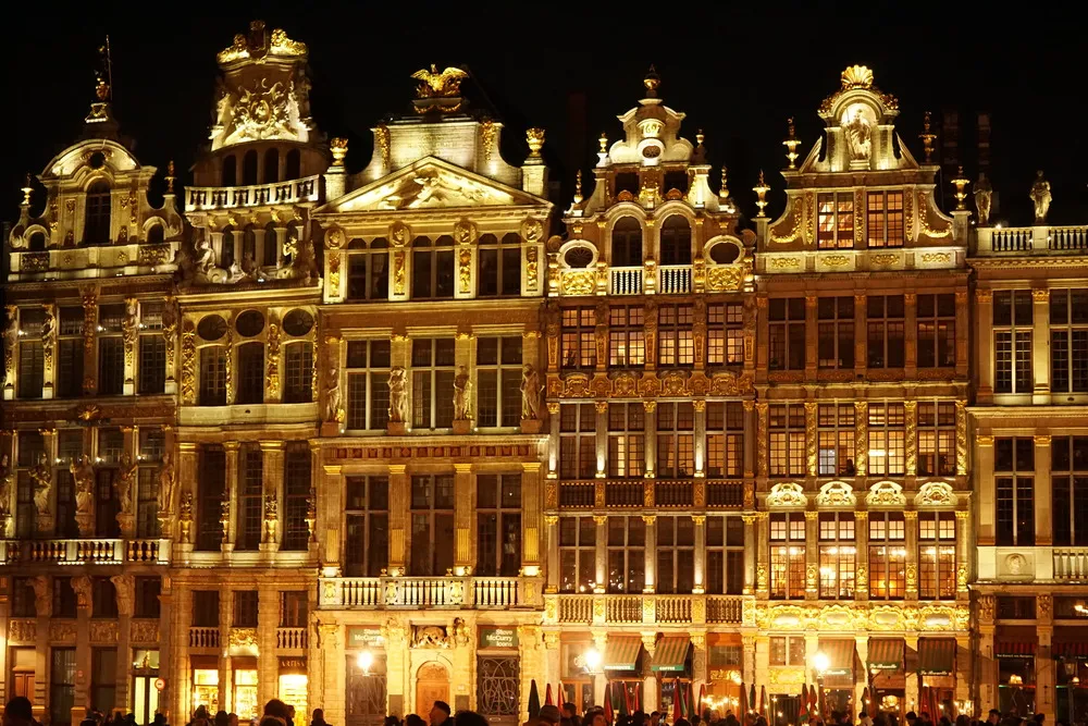 Gelb beleuchtete Fassade von Altstadtgebäuden mit zahlreichen Verzierungen in der Nacht. An den Giebeln befinden sich teilweise goldene Skulpturen.