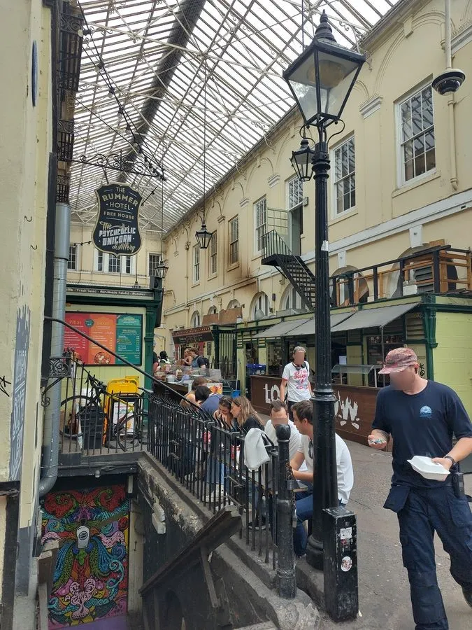 Eine Gasse mit Glasdach und Geschäften links und rechts. Eine Treppe im Vordergrund führt in einen Clubraum.