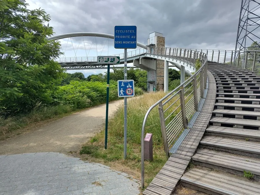 Fußgänger- und Radfahrerbrücke bei Nanterre. Breite Holzstufen führen über einen breiten Bogen auf die Brücke, an der linken Seite gibt es eine schmale Rampe für Radfahrer