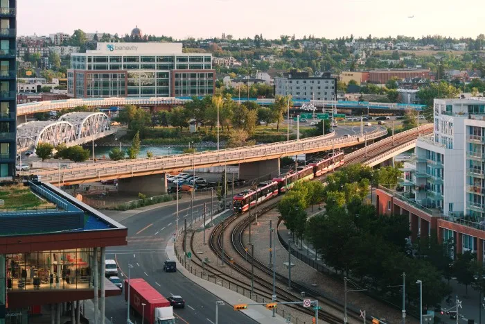 Blick aus der Vogelperspektive auf eine belebte Stadtlandschaft. In der Mitte des Bildes fährt eine Straßenbahn, daneben fahren einige Autos. Die Straßenbahnschienen hinter der Straßenbahn überqueren einen Fluss. Vier weitere Brücken für Autos führen über denselben Fluss auf der rechten Seite. Im Hintergrund befindet sich ein Hügel mit kleineren Gebäuden und mehr Grün. Am Himmel ist eine Ebene zu sehen. Im Vordergrund links ist ein Wolkenkratzer kaum zu erkennen.