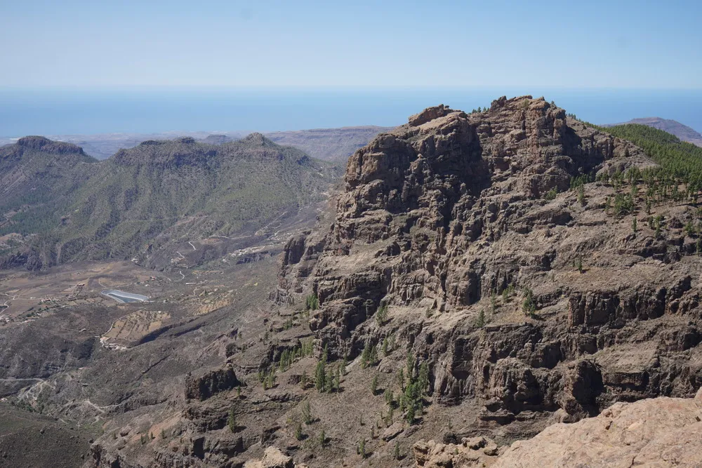 Weiter Blick hinab in eine trockene, steinige Landschaft. Im Hintergrund das Meer. Rechts im Bild in weiter Ferne ein kleiner Nadelwald.