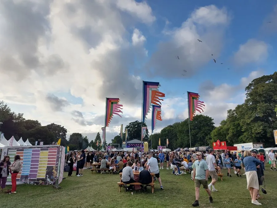Auf einer grünen Wiese wurden zahlreiche Zelte und bunte Banner aufgestellt. Auf Sitzbänken und um sie herum befinden sich zahlreiche Menschen.