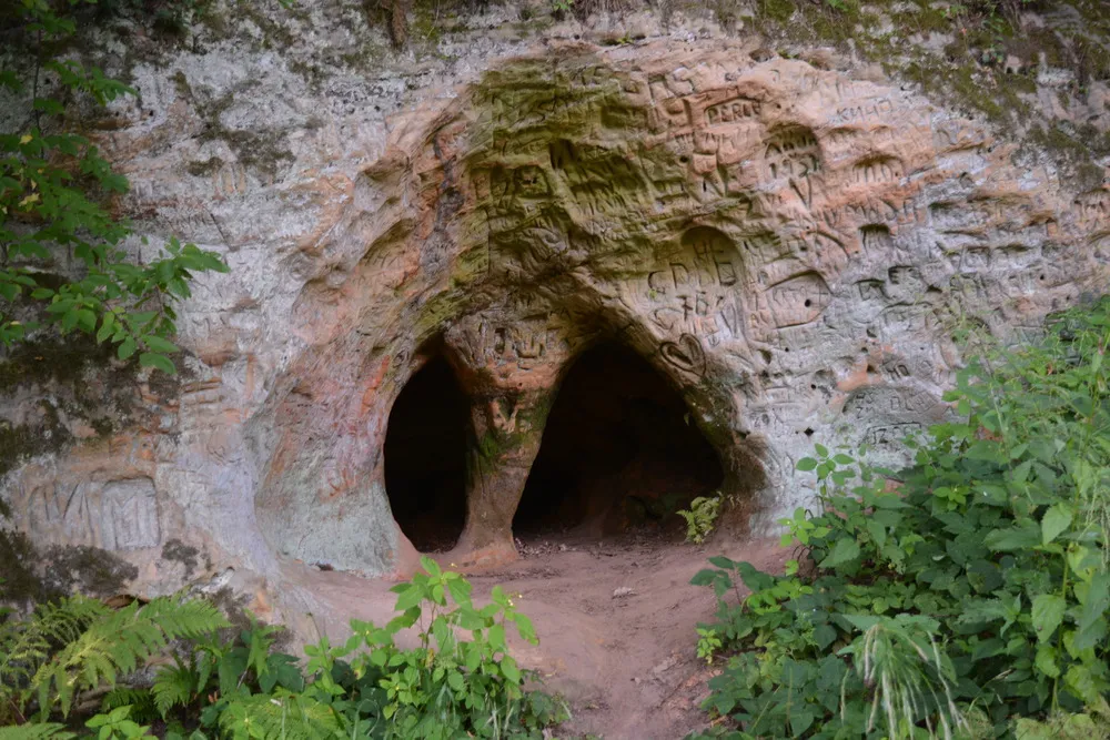 Höhleneingang in Sandstein mit zahlreichen Gravierungen. In der Mitte des Eingangs ist eine natürliche Säule.