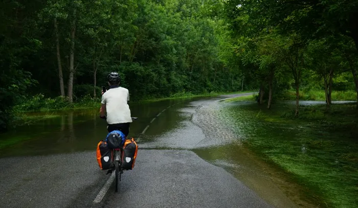 Überflutete Straße mitten im Wald, die einem Radfahrer im Vordergrund den Weg versperrt.