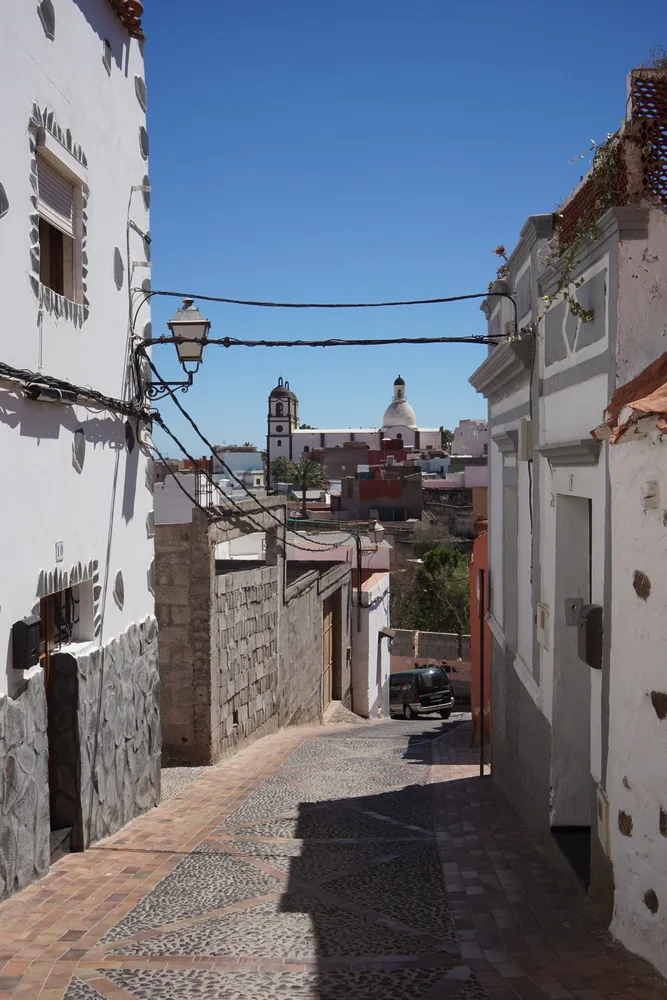 Eine schmale gepflasterte Straße führt bergab. Daneben stehen enge aneinander alte Häuser mit weißer Fassade. Im Hintergrund steht eine Kirche auf einem Berg.