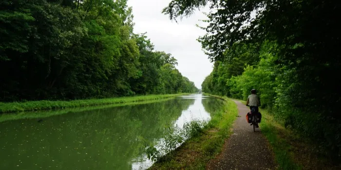 Ein einsamer Radfahrer fährt auf geteertem Radweg links eines geradlinigen Kanals umgeben von Bäumen.