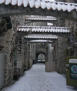 Verschneite mittelalterliche Gasse. Wände aus Mauersteinen links und rechts, welche mit geziegelten Bögen verbunden sind. Am Ende der Gasse ist ein Tor.