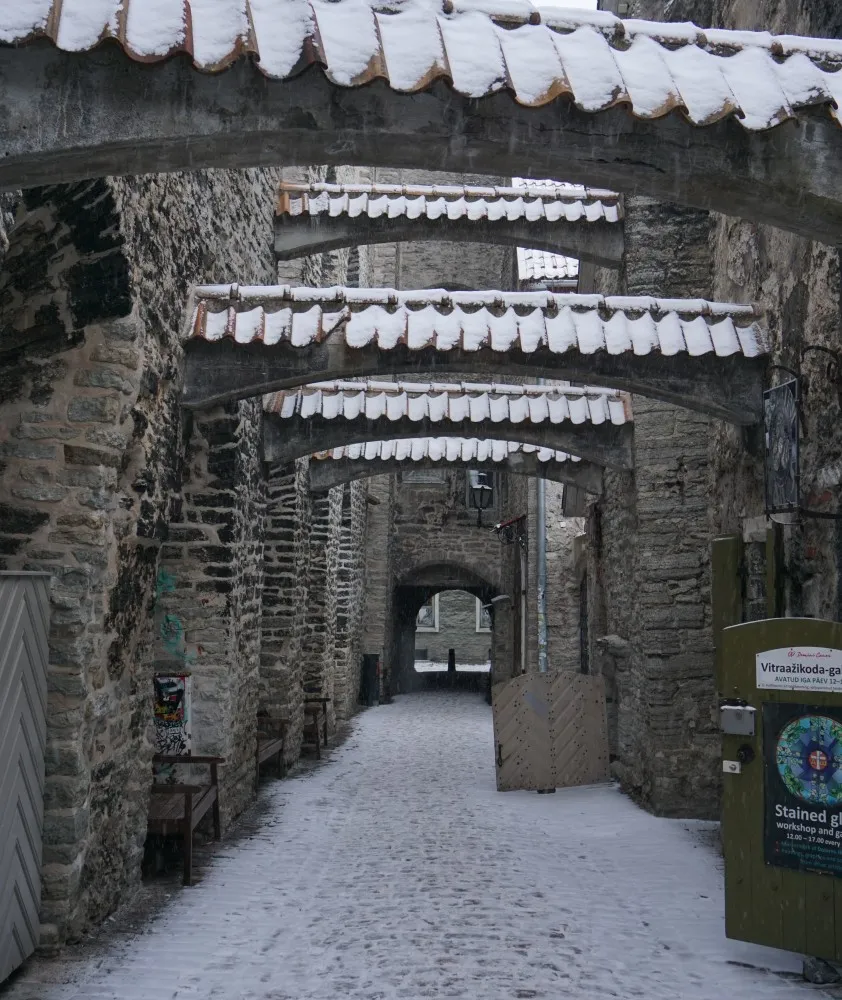 Verschneite mittelalterliche Gasse.
Wände aus Mauersteinen links und rechts,
welche mit geziegelten Bögen verbunden sind.
Am Ende der Gasse ist ein Tor.