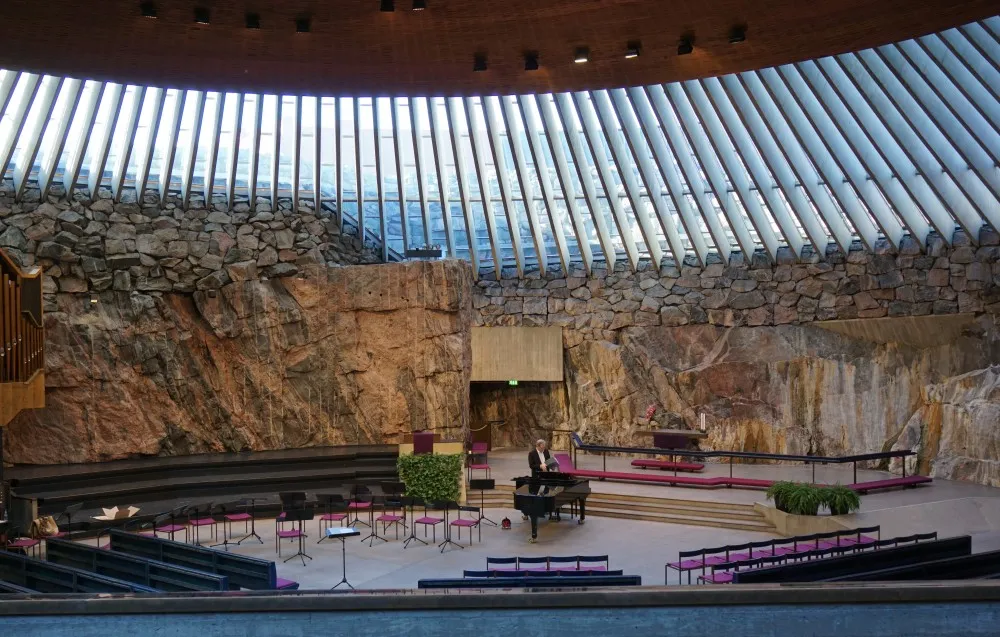 Das innere eines runden Kirchengebäudes. Das Dach ist eine moderne Holz- und Glaskonstruktion. Die Wände bestehen aus natürlichem Felsen. In der mitte spielt jemand auf einem Flügel.
