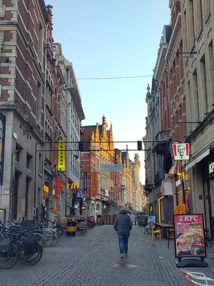 Rotes Sonnenlicht bestrahlt die Altstadtfassade in einer Fußgängerzone. An den alten Häusern hängt viel Leuchtreklame für McDonalds, KFC und weiteren Läden.