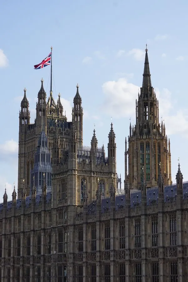 Die Frontfassade und einige Türme des Buckingham Palace. Heller, warmer Stein, zahlreiche Türmchen und Fensternischen sind zu sehen. Auf einem der Türme weht die Flagge von Großbritannien.