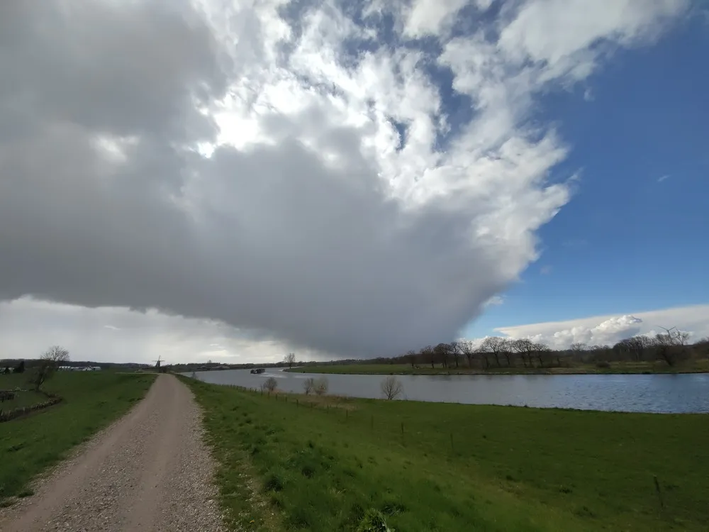 Eine große dunkle Regenfront zieht über einen Fluss. Davor blauer Himmel. Im Vordergrund führt ein Schotterweg neben dem Fluss auf einem grün bewachsenen Deich nach hinten zu einer Windmühle.