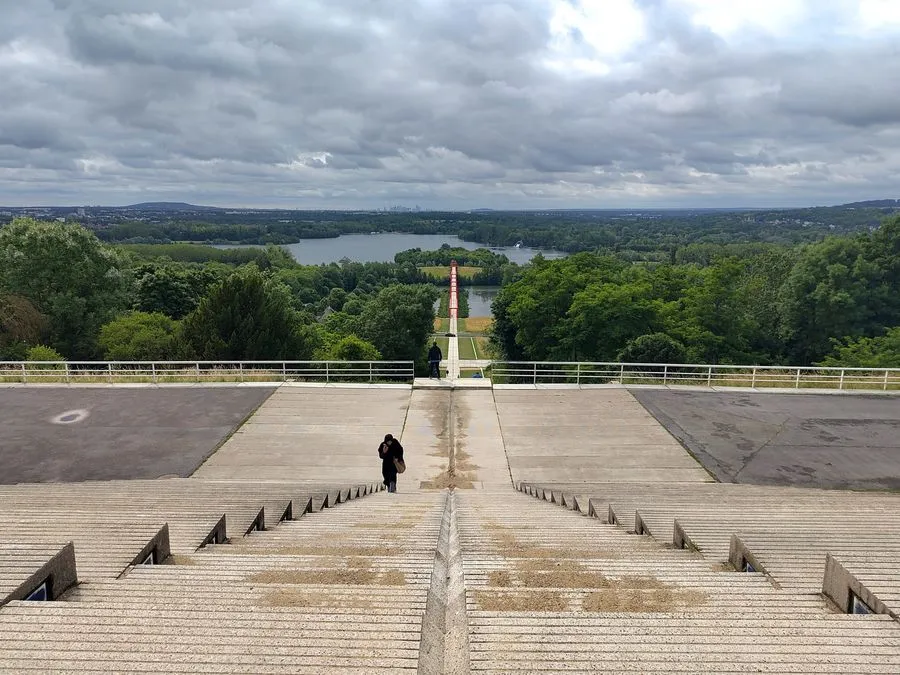 Treppen führen mittig im Bild von einer Anhöhe in die tiefe des Bildes. Dort befindet sich eine Parkanlage und ein See mit Insel. Am Horizont ist die moderne Skyline von Paris zu sehen.