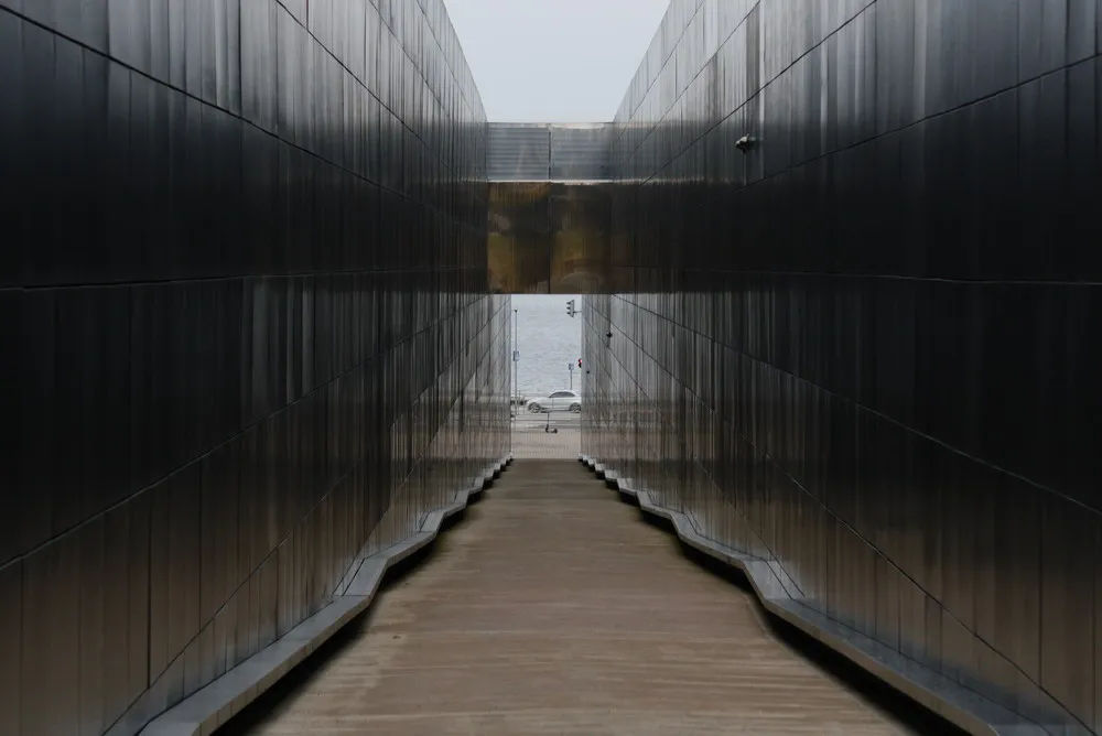 Ein dunkler Gang führt ans Meer. Links und rechts schwarze Wände mit zahlreichen eingravierten Namen.