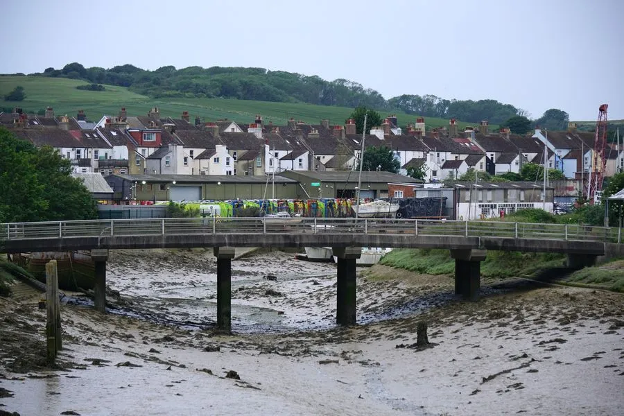 Ein trockenes Flussbett kommt aus einer britischen Siedlung in den Vordergrund. Eine kleine Fußgängerbrücke überquert es. Im Hintergrund ist eine hügelige Wiesenlandschaft zu sehen.