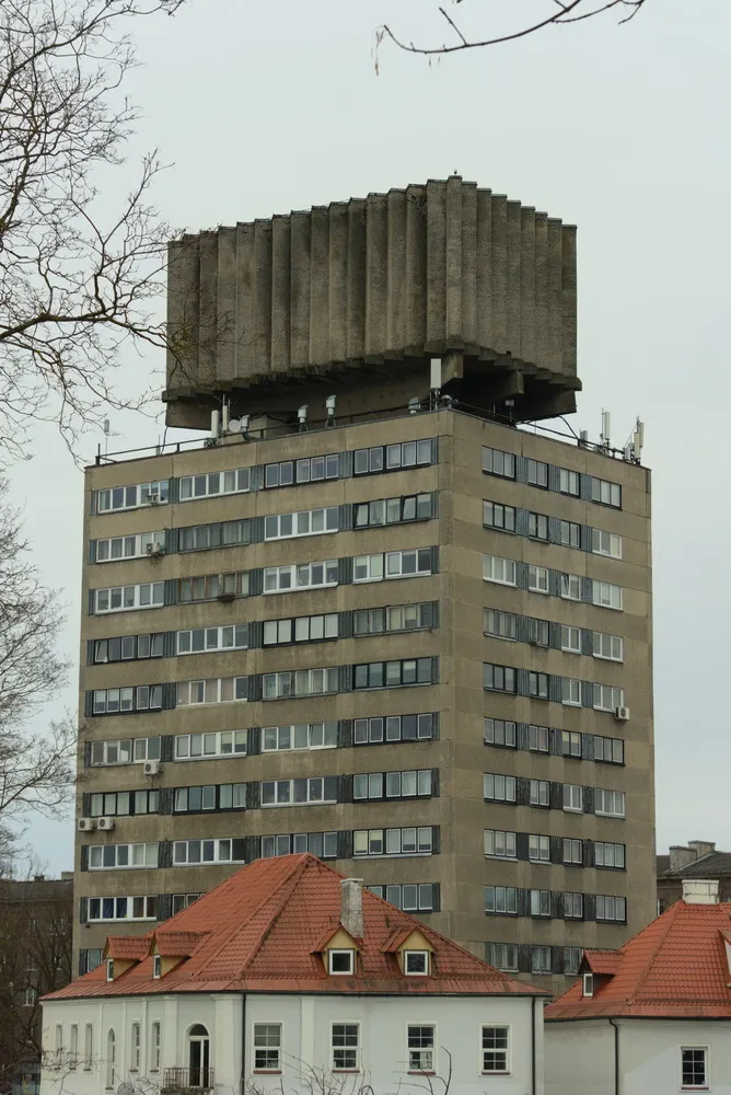 Ein großes, graußes Betongebäude mit simpelster Fassade streckt sich in den Himmel. Auf dem Dach des Blocks steht ein gigantischer Betonwürfel mit einer Art Akkordeonstruktur, vermutlich ein Kunstwerk. Zwischen Block und Dach befinden sich zahlreiche Antennen.