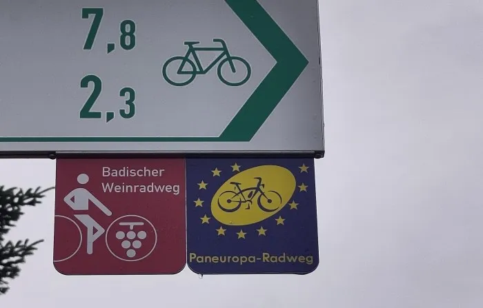 Das Schild des Paneuropa-Radwegs hängt an einem deutschen weiß-grünen Radwegweiser. Das Logo is ein blaues Rad auf einer gelben Ellipse umgeben von Sternen.