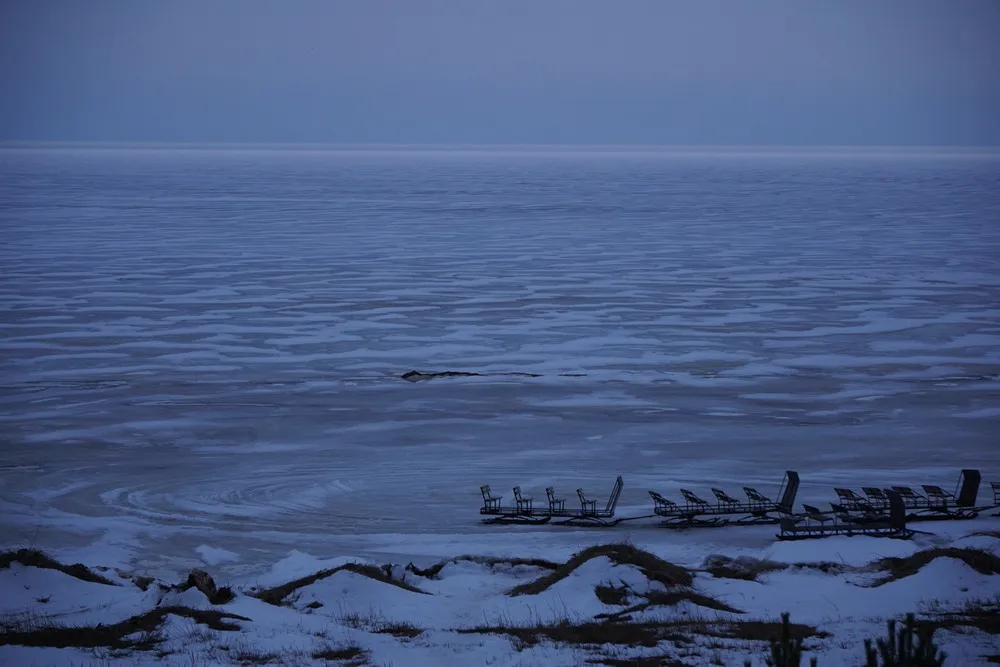 Lila Abenddämmerung über einem gigantischen, eingefrorenem See. Am Ufer des Sees befinden sich 4 große Schlitten.
