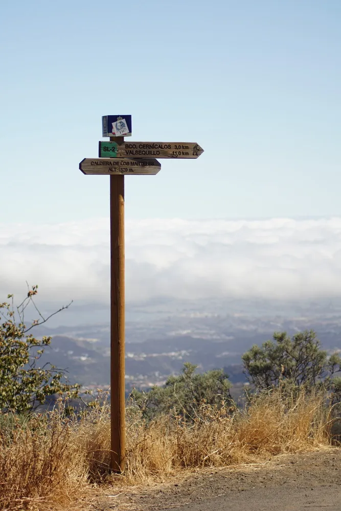 Ein Schild weist eine Wanderroute am Straßenrand aus. Im Hintergrund sieht man tief hinab bis an die Küste der Insel. Man sieht ebenfalls auf Wolken hinab.