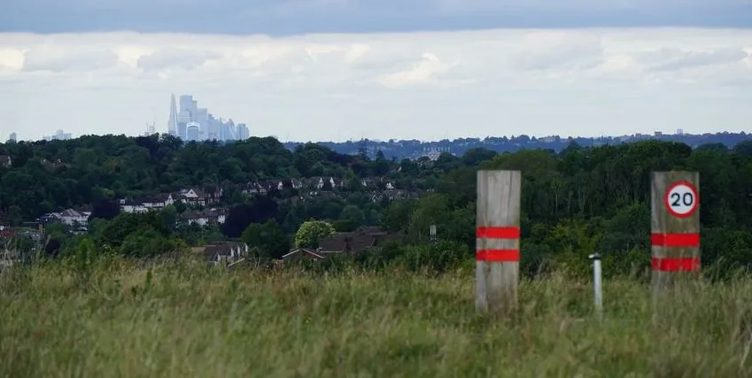 Die Skyline von London baut sich am Horizont auf. Im Vordergrund sind Holzbalken mit einem Tempo-20 Schild (Meilen pro Stunde), die die Straße auf dem Red Hill markieren.