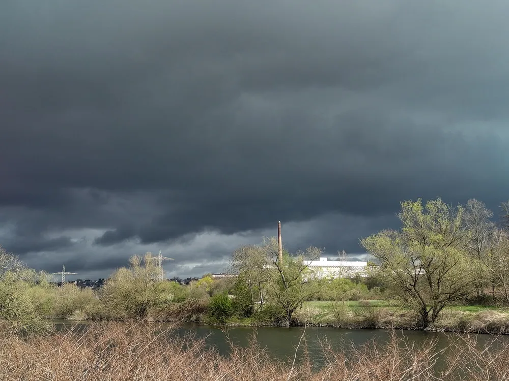 Dunkelgraue Gewitterwolken über einem Fabrikschornstein. Im Vordergrund Bäume und ein Fluss, die Ruhr.