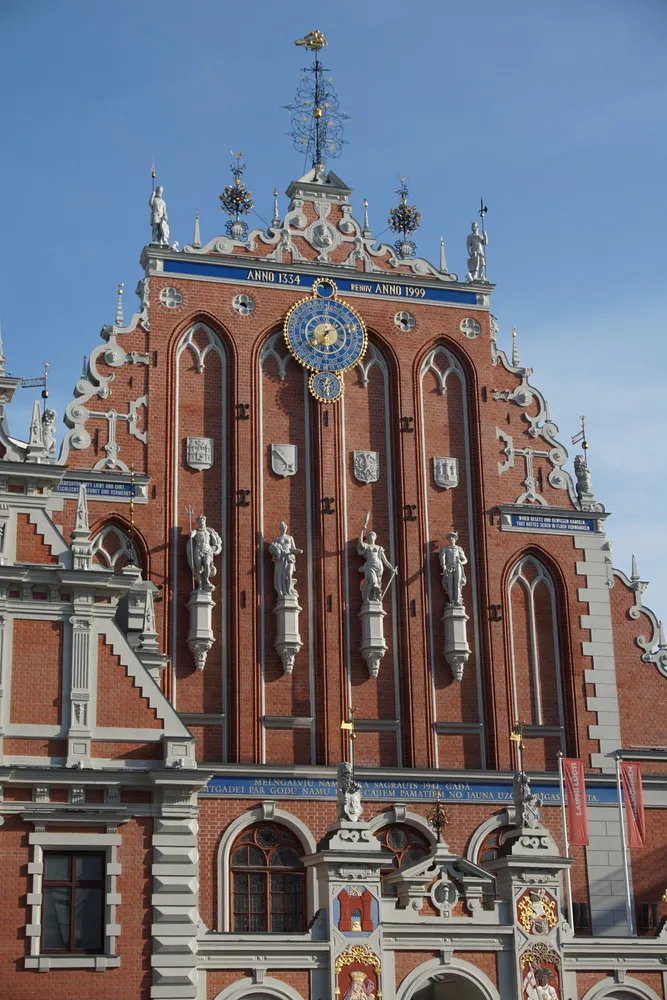 Fassade eines Ziegelbaus mit zahlreichen kunstvollen Verzierungen durch Statuen, Wappen und weiterem. Oben in der Mitte eine blau-goldene Uhr.