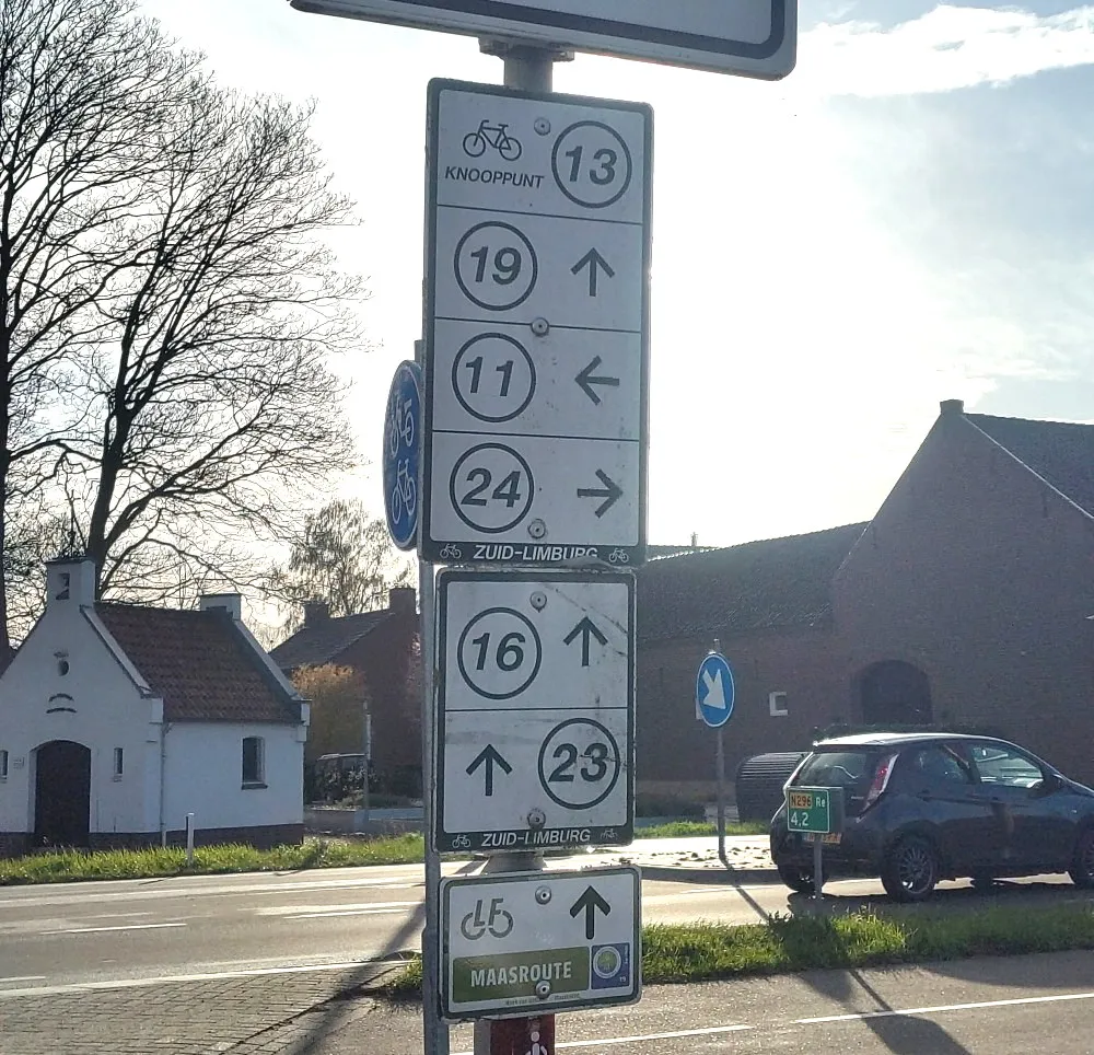 Schild an einer Straße. Zu sehen sind Wegweiser für 6 verschiedene Knotenpunkte und ein Schild für die Maasroute.
