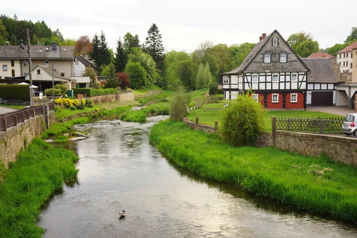 Blick von einer Brücke über der Spree nach Schirgiswalde. Grünes Ufer, rechts ein Fachwerkhaus, im Hintergrund Wald. Eine Ente sitzt in der Spree.
