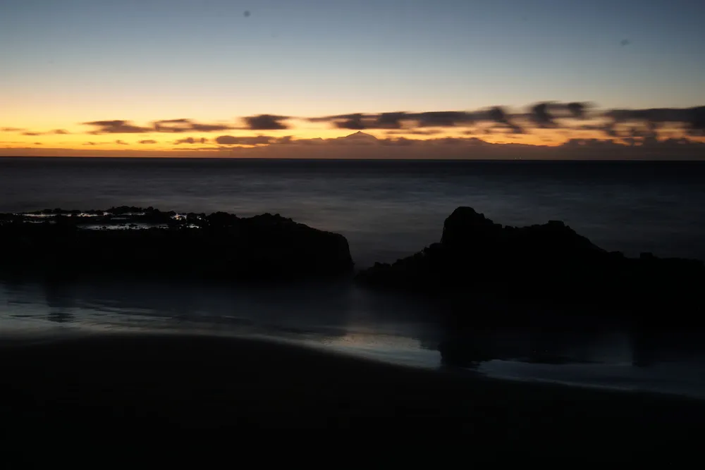 Die Sonne geht hinter der Insel Teneriffa unter, welche am Horizont zu sehen ist. Wolken verdecken die Insel teilweise.