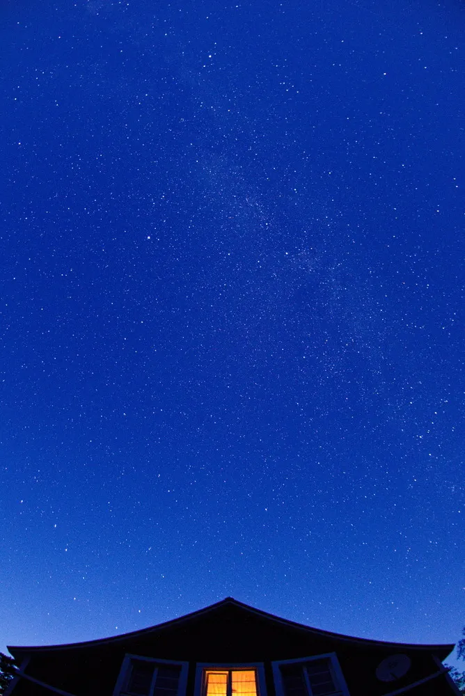Blauer, klarer Sternenhimmel über einem roten Holzhäuschen mit gelb leuchtendem Fenster.