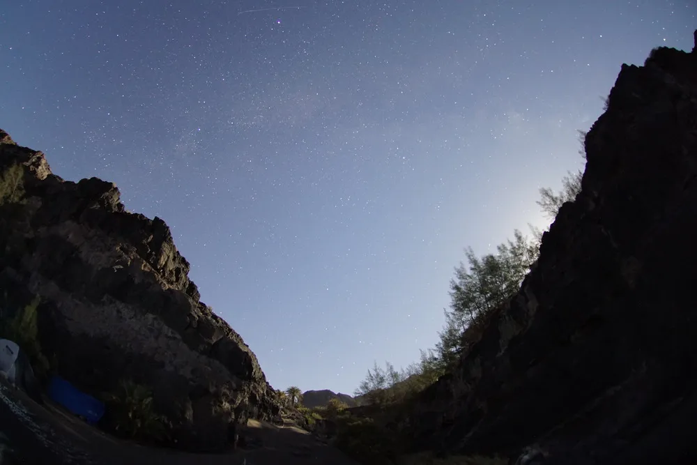 Die Milchstraße erstreckt sich über eine Tal. Das Foto ist durch das Fisheye Objektiv stark verzerrt. Rechts leuchtet der Mond hinter den Bergen hervor und beleuchtet die linke Talseite.