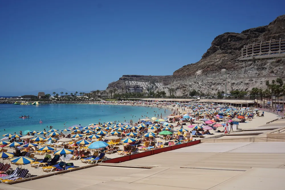 Ein Strand voller Liegen und Sonnenschirme. Im Hintergrund erhebt sich ein Berg, an dem ein riesiges modernes Hotel gebaut wurde. Links leuchtet das blau-türkise Wasser. Blauer Himmel ohne Wolken.