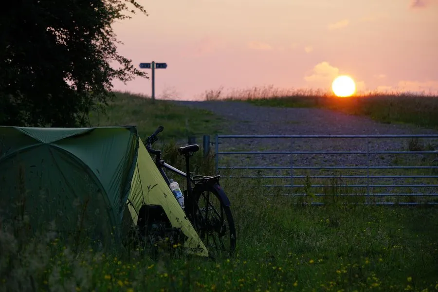 Die rote Sonne geht unter. Im Vordergrund steht ein grünes Zelt und ein Fahrrad. Links neben der Sonne steht ein Wegweiser.