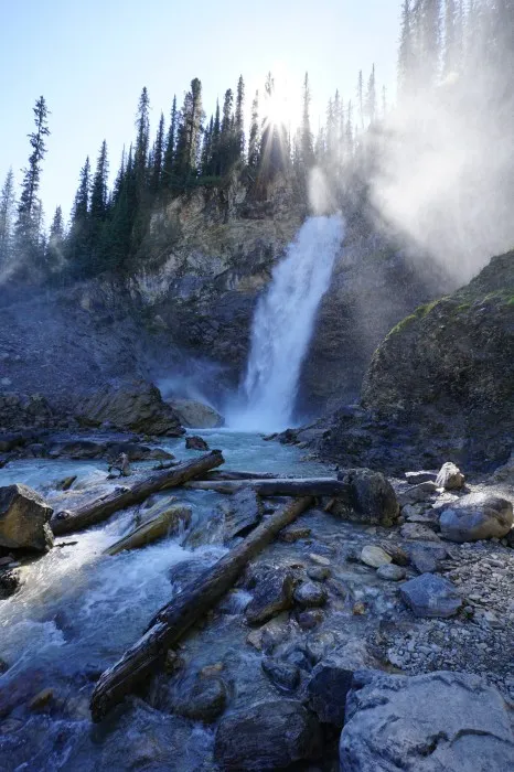 Ein 30 Meter hoher Wasserfall, der einen Felsen hinabstürzt. Oben auf den Felsen ist ein Nadelwald, unten ein wilder Back. Die Sonne ragt gerade so aus dem Wald auf dem Felsen hervor, ihre Strahlen sind durch das aufgesprühte Wasser sichtbar.
