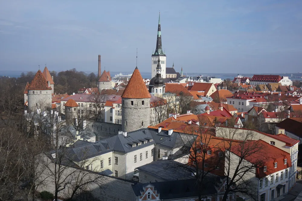 Ausblick von oben auf die Stadt Tallinn. Die Sonne scheint, und zahlreiche Türme von Stadtmauer und Kirchen sind zu sehen.