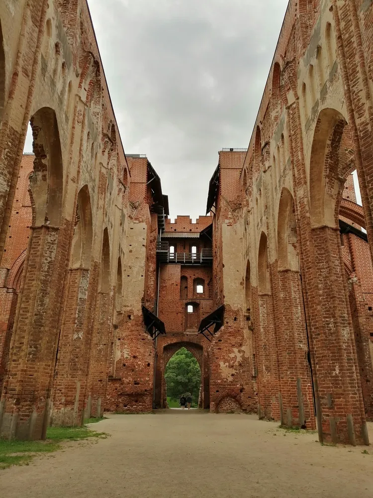 Ruine einer Kirche aus rotem Ziegel. Das Dach fehlt, der Boden ist Sand und Wiese. Die Wände stehen teilweise noch.