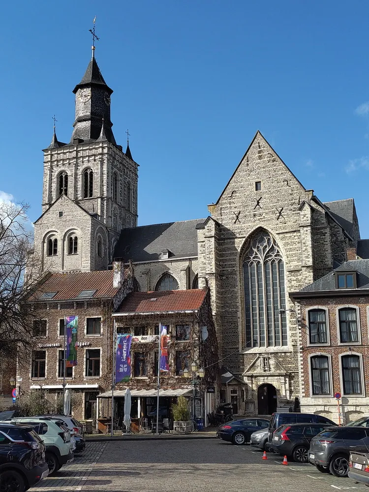 Alte Kirche aus Stein mit einem Turm links und einem großen Fenster rechts. Davor ein mit Efeu bewachsenes Haus und Autos auf Parkplätzen.
