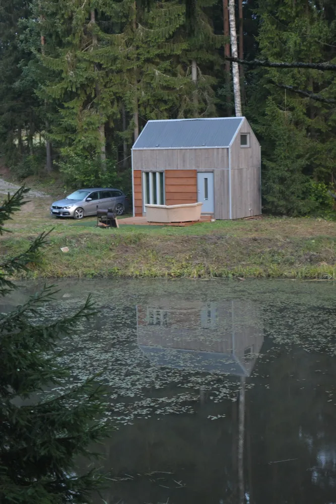 Ein kleines, hölzernes, modern gebautes Haus mit Metallschienen und Blechdach. Daneben ein silbernes Auto. Im Vordergrund ein See, in dem sich das Haus spiegelt.