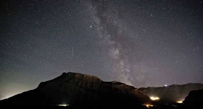Klarer Nachthimmel über einer dunklen Gebigsfront. In dem dunklen Wald vor dem Gebirge können einzelne Lichtquellen von Häusern ausgemacht werden.