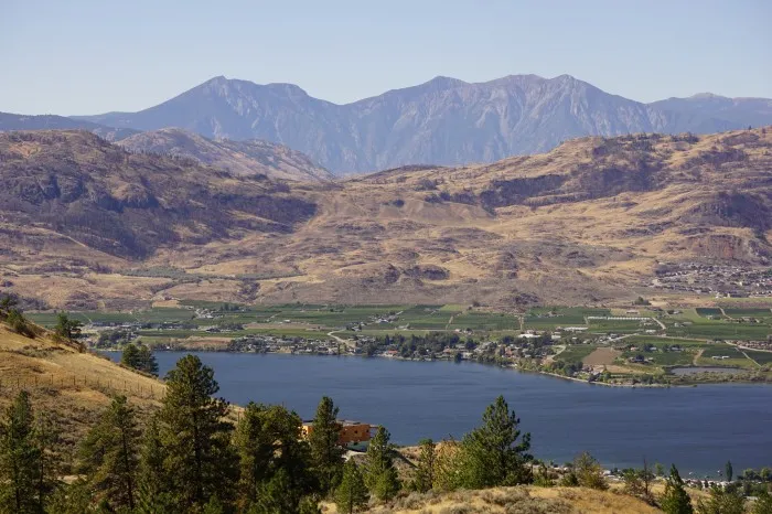 Berge aus trockenem, orange-gelben Fels türmen sich hinter einem See auf. Am Ufer des Sees sind einige grüne Felder zu sehen. Im Vordergrund endet ein Nadelwald.