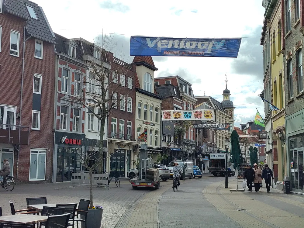 Eine kleine Straße führt durch eine Innenstadt mit braunen und gelben Gebäuden. Über der Straße hängt ein blaues Banner.