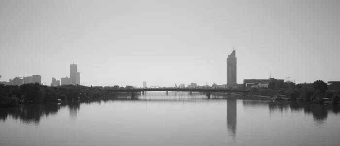 Schwarz-Weiß Foto der Donau in Wien. Links und rechts sind einige Bäume und Hochhäuser zu sehen. In der Mitte ein paar Brücken.