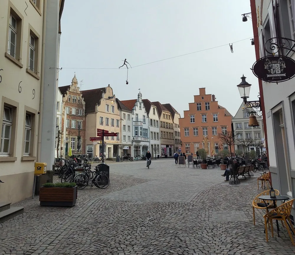 Bunte Altstadthäuser auf einem gepflasterten Marktplatz. Eine Skulptur eines Seiltänzers steht auf einem darübergespannten Seil.