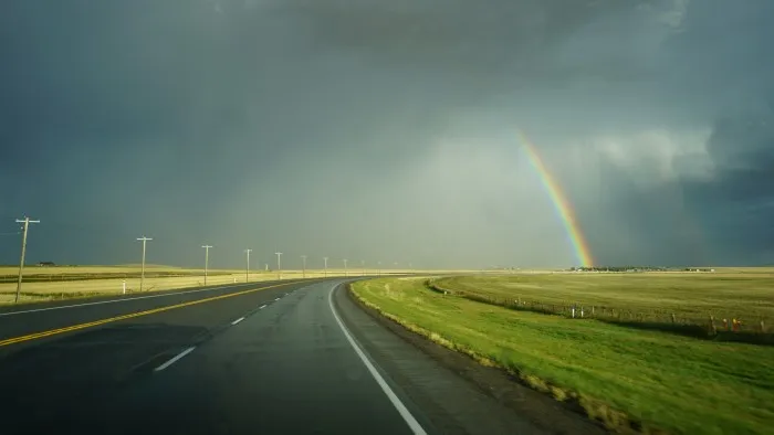 Ein doppelter Regenbogen strahlt vor einer dunkelgrauen Wolkenfront über einem gelb-grünem Feld. In der Mitte des Weges geht eine geteerte Straße in eine Rechtskurve über. Ein Strommast begleitet sie auf der linken Seite.