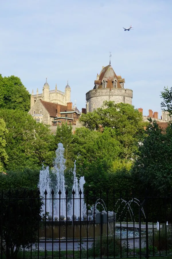 Die Burg Windsor baut sich auf einem Hügel hinter einer grünen Parkanlage mit Springbrunnen auf. Ein Flugzeug überfliegt die Szene.